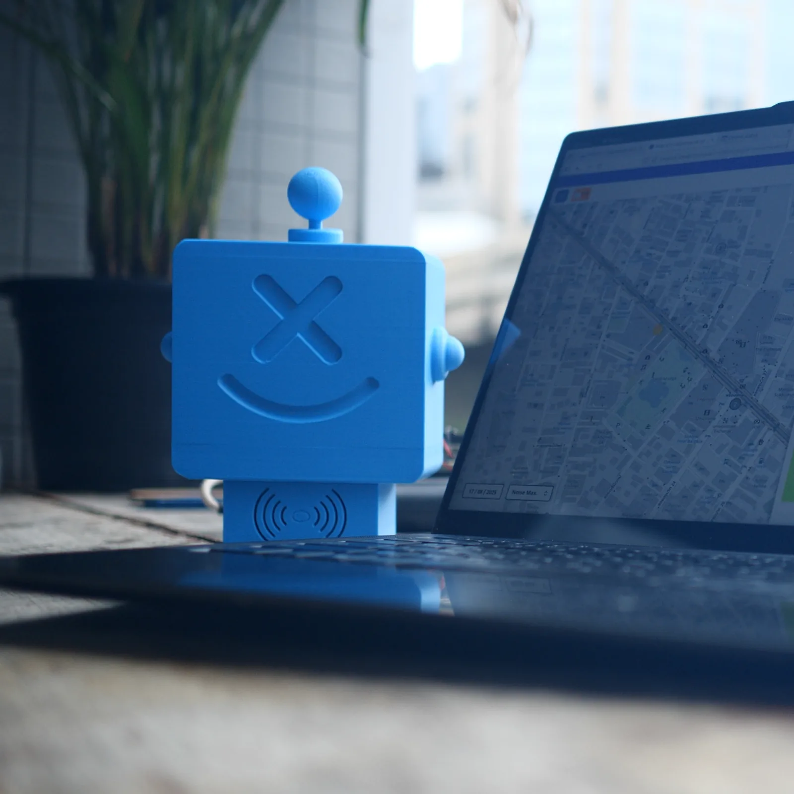 A blue Altruist on a desk beside an open laptop showing the sensors.social map